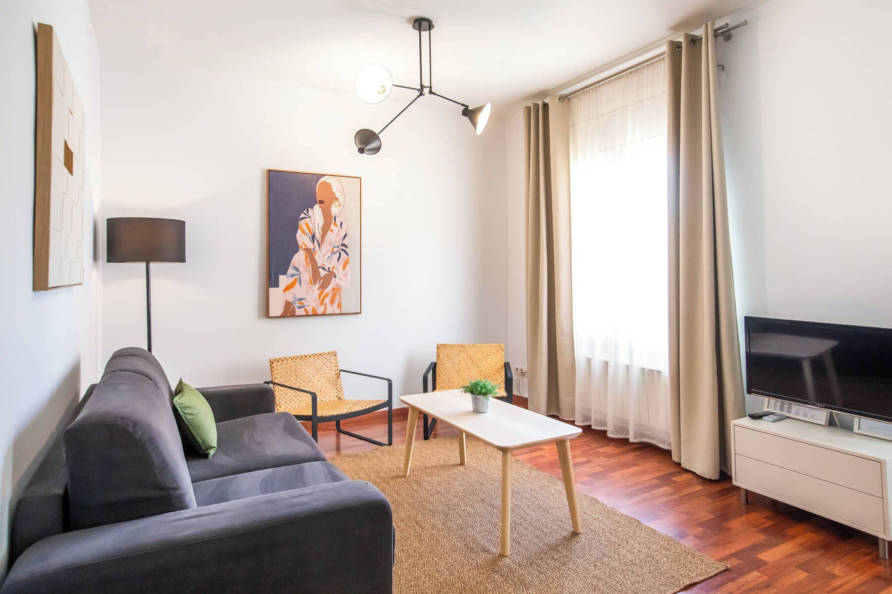 A modern living room with a grey sofa, two wicker chairs, a small white table, a floor lamp, a painting, a TV, and a window with curtains.