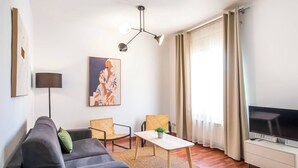 Ein modernes Wohnzimmer mit grauem Sofa, zwei Korbstühlen, einem kleinen weißen Tisch, einer Stehlampe, einem Gemälde, einem Fernseher und einem Fenster mit Vorhängen.
