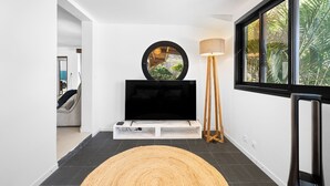 Una sala de estar moderna con un televisor de pantalla plana sobre un soporte blanco, un espejo redondo, una lámpara de pie y un ventanal con vistas a zonas verdes.