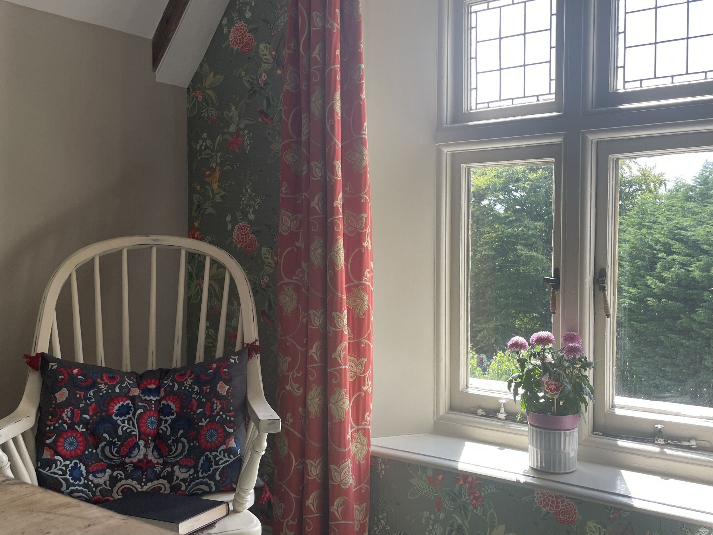 A room with a white chair, floral wallpaper, a window with a view of greenery, and a potted plant on the windowsill.