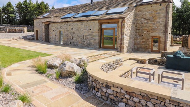 A stone building with a sloped roof, large windows, and a stone pathway leading to a seating area. - Carden Laithe Barn (Skipton)