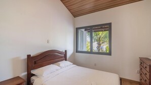 A bedroom with a wooden headboard, a single bed, a nightstand, a dresser, and a window with a view of palm trees. - House in Cabo Frio 200m From the Beach Adp001 (Cabo Frio)