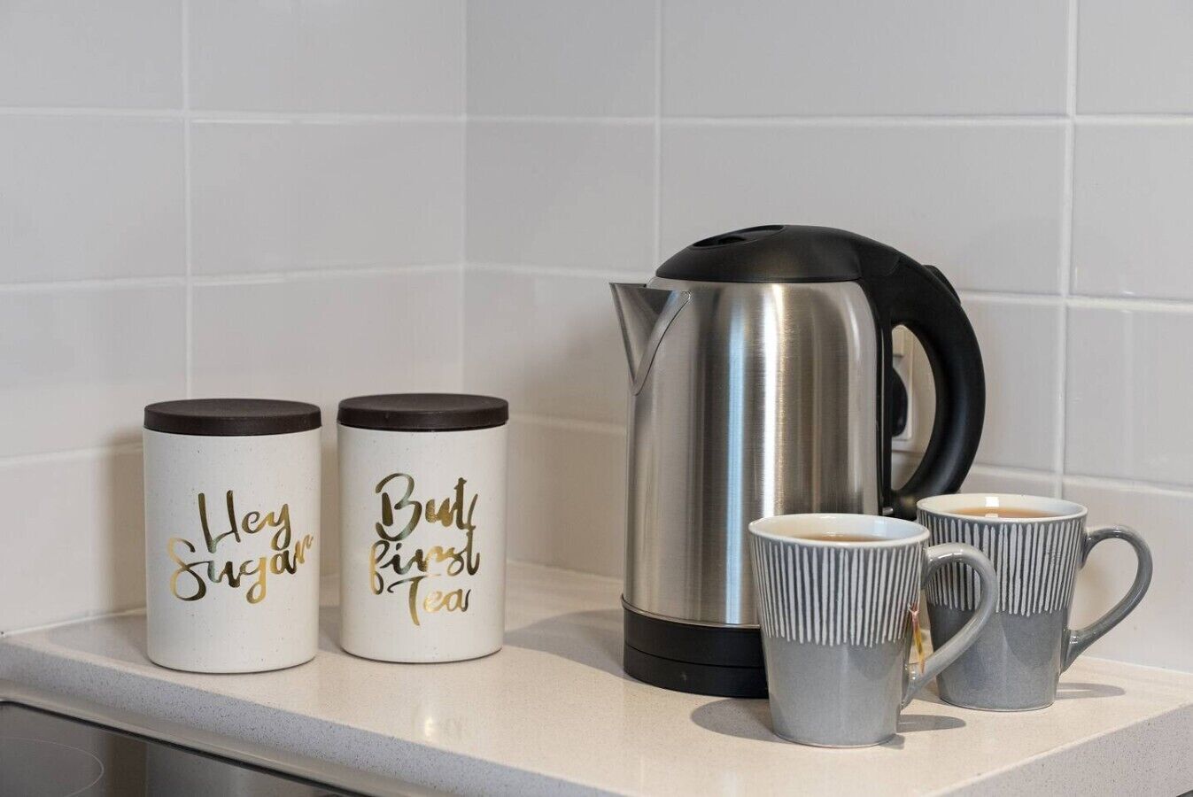 A kitchen counter with two white mugs with gold text, a silver kettle, and two gray ribbed mugs.