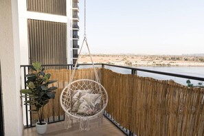A balcony with a hanging chair, a potted plant, and a view of a beach and cityscape.