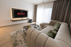 A modern living room with a large flat-screen TV mounted on the wall, a beige sofa, a coffee table with fruits, and a patterned rug.