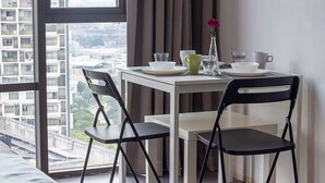 Ein modernes Zimmer mit Esstisch, zwei Stühlen, einer Vase mit einer Blume und Blick auf eine Stadtlandschaft durch große Fenster.