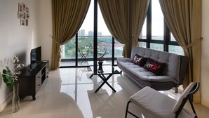Ein modernes Wohnzimmer mit einem grauen Sofa, einem Fernseher auf einem Schrank, einem Glastisch und Blick auf eine Stadtlandschaft durch große Fenster mit beigen Vorhängen.