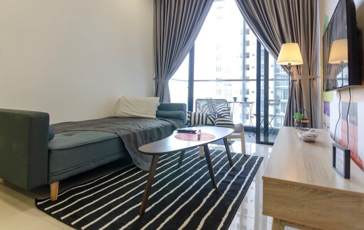 Una habitación de hotel moderna con sofá, una mesita redonda pequeña, un escritorio de madera y un ventanal con cortinas.
