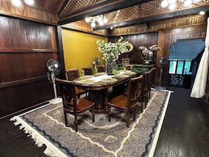 Un coin repas avec une table en bois dressée pour six personnes, entourée de chaises, d’un tapis à motifs et d’un ventilateur.