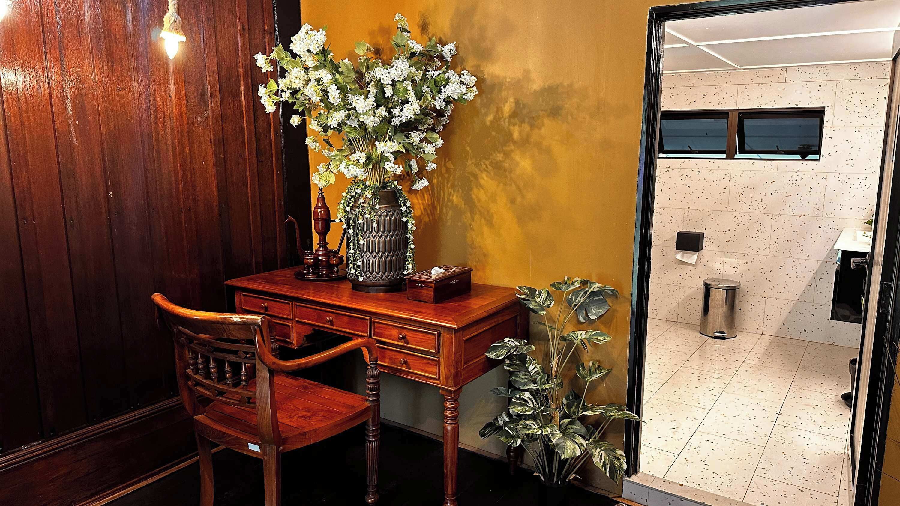 Ein Holztisch mit einer Vase weißer Blumen, ein Stuhl und ein Blumentopf in einem Raum mit einer Holzverkleidung und Fliesenboden.