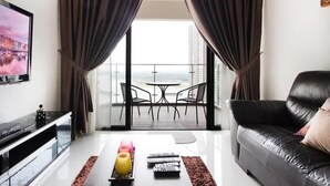 A modern living room with a black leather sofa, a glass coffee table with remote controls, a TV on the left, and a balcony with a table and chairs. - High-floor 2BR Suite, Water Views, Free Parking (Johor Bahru)