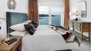 A hotel room with a large bed, a wooden nightstand, a desk, a mirror, and a view of the sea.