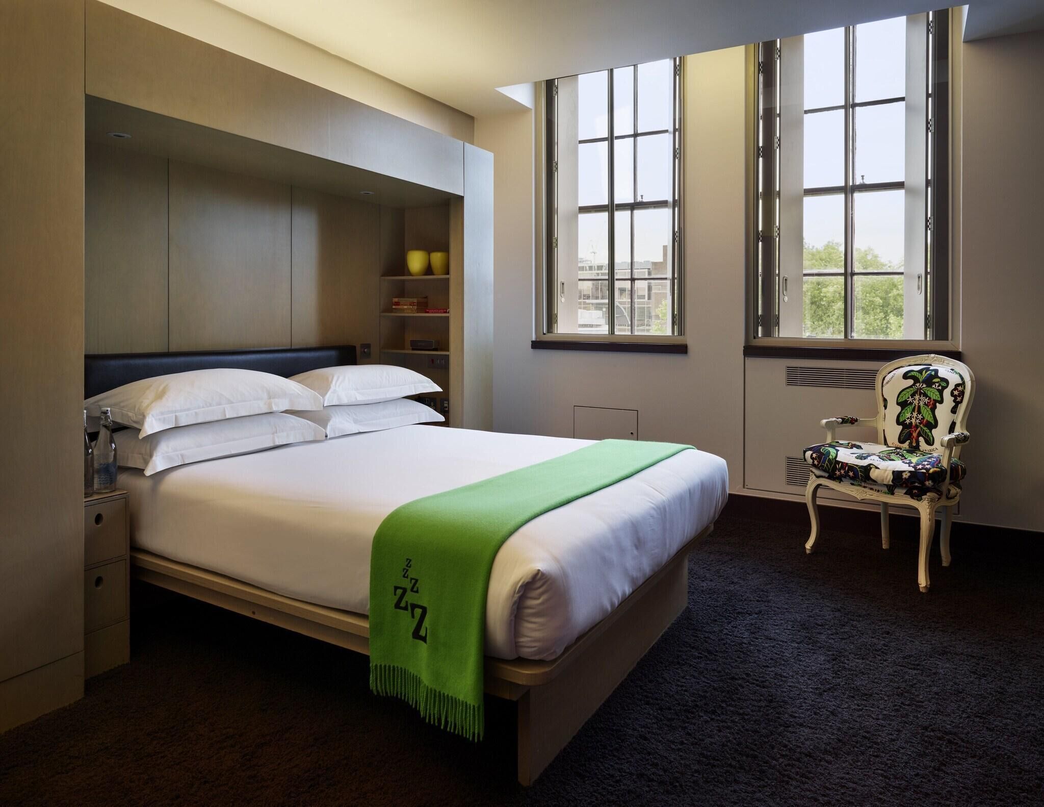 A neatly made bed with a green blanket, a chair with floral upholstery, and a large window with a view of buildings.