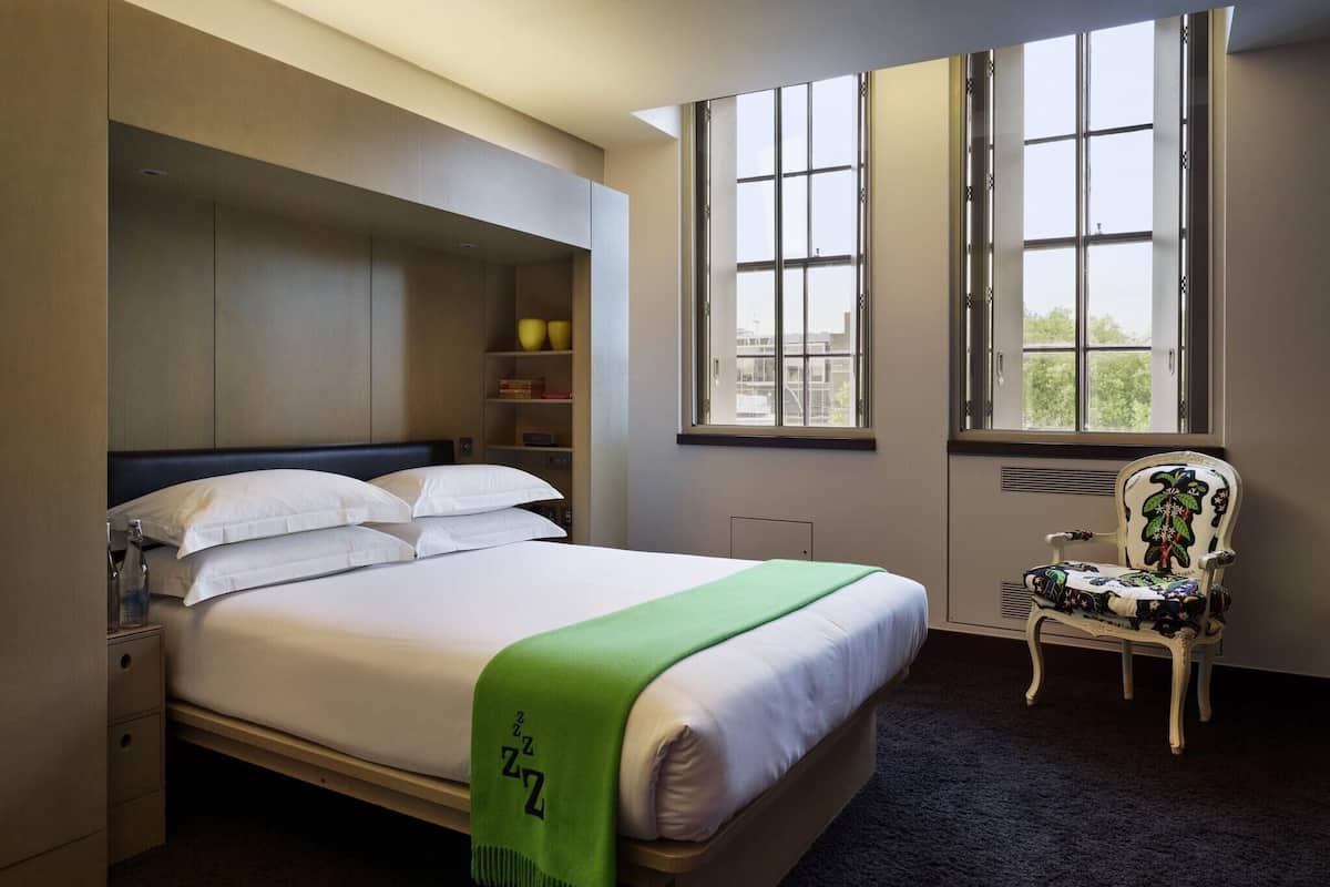 A neatly made bed with a green blanket, a chair with floral upholstery, and a large window with a view of buildings.