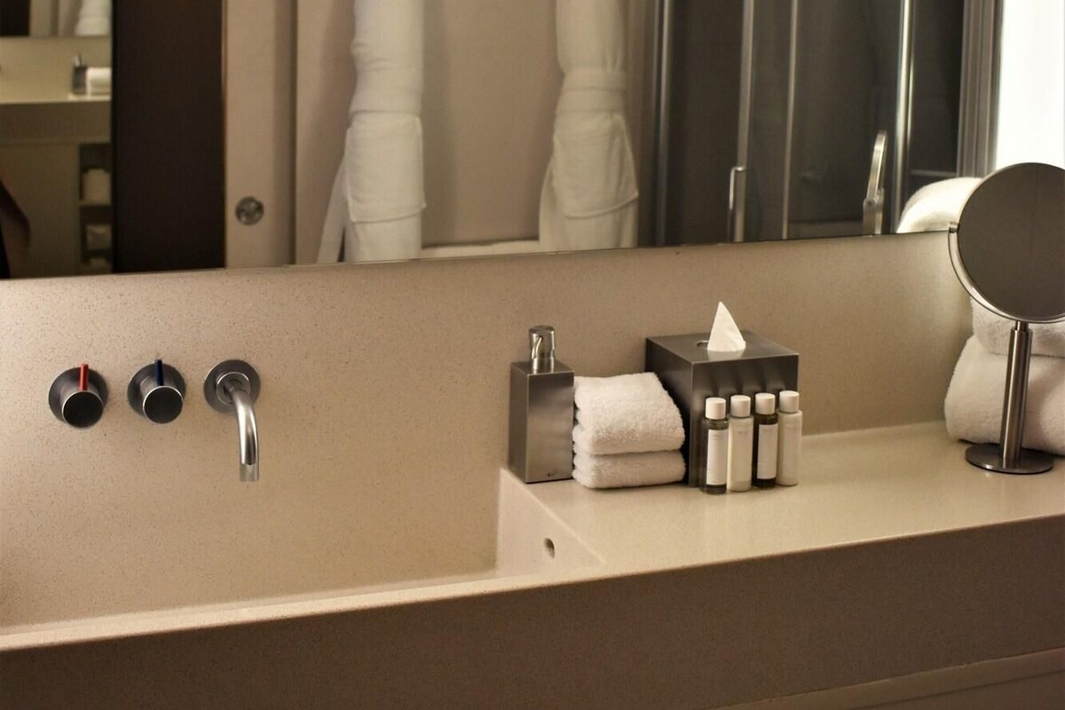 A modern bathroom with a large mirror, a sink with three faucets, and a towel rack with two white towels.
