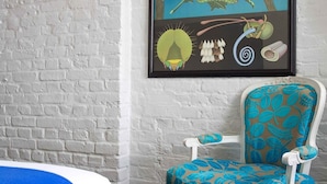 A room with a white brick wall, a framed butterfly and caterpillar illustration, a blue and white patterned chair, and a blue blanket with "ZZ" on it. - The Zetter (London)