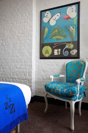 A room with a white brick wall, a framed butterfly and caterpillar illustration, a blue and white patterned chair, and a blue blanket with "ZZ" on it. - The Zetter (London)