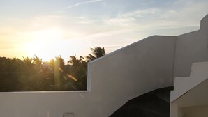 A modern staircase with a white exterior and black steps, leading to a rooftop area with a clear view of the sunset and palm trees in the background.
