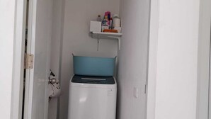 A small, narrow room with a white wall, a white door, and a white air conditioning unit. There is a blue cushion on the floor and a white shelf with various items on it.