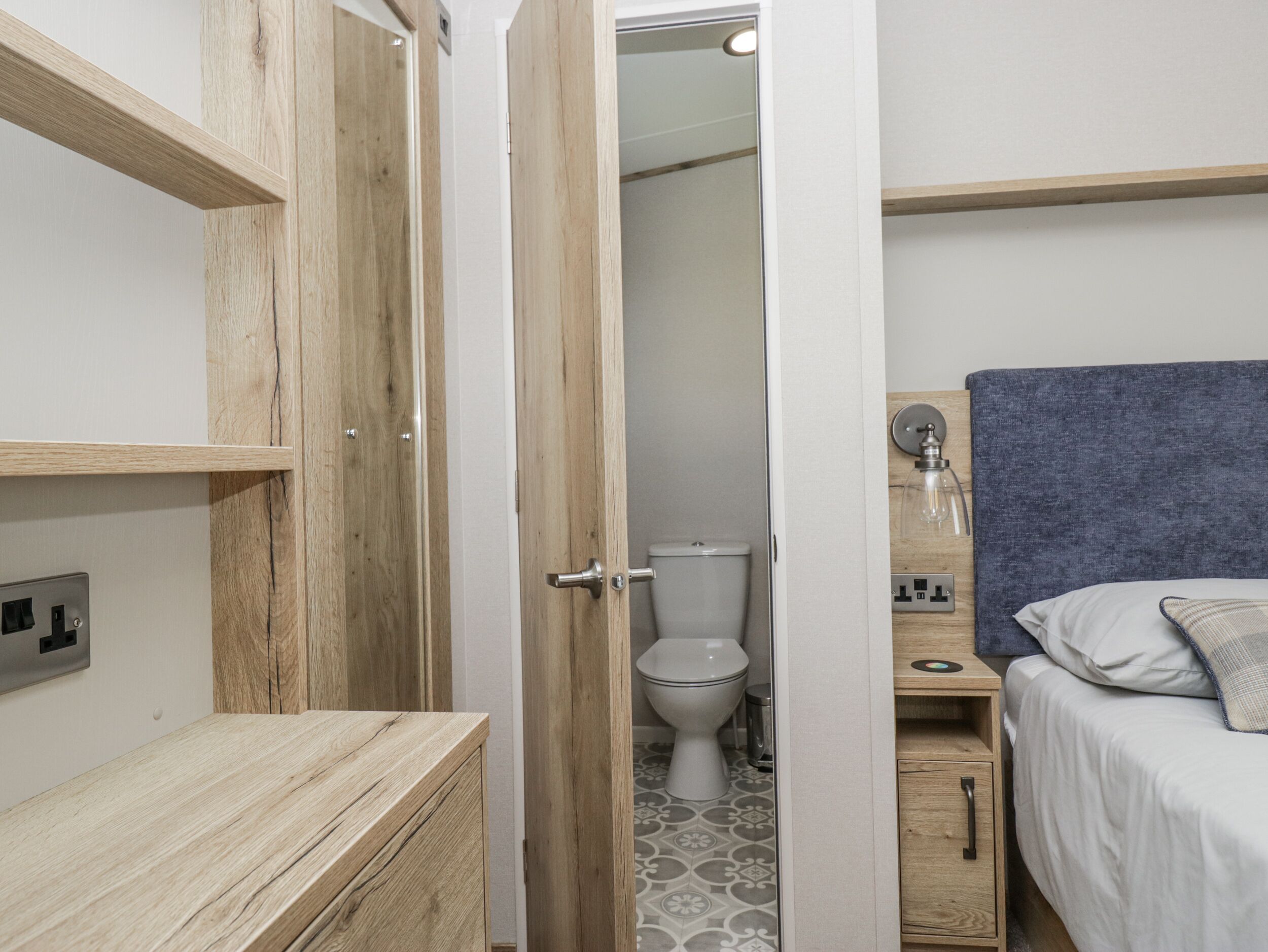 A small, compact bedroom with a wooden wardrobe, a bedside table, a bed with a headboard, and a bathroom with a toilet and sink.