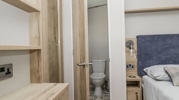 A small, compact bedroom with a wooden wardrobe, a bedside table, a bed with a headboard, and a bathroom with a toilet and sink.