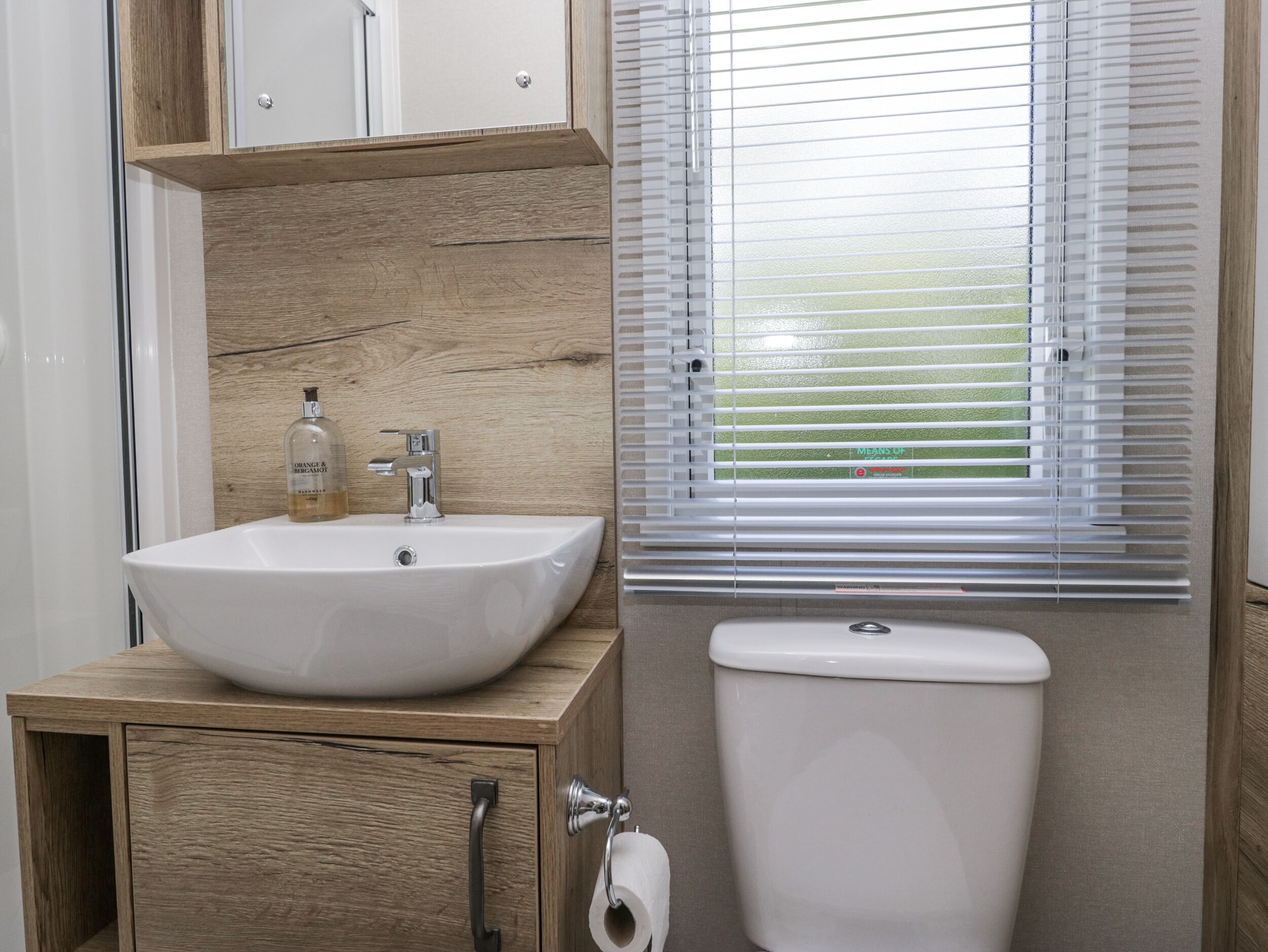 A bathroom with a wooden vanity, a white sink, a bottle of orange & bergamot body wash, a toilet, and a window with blinds.