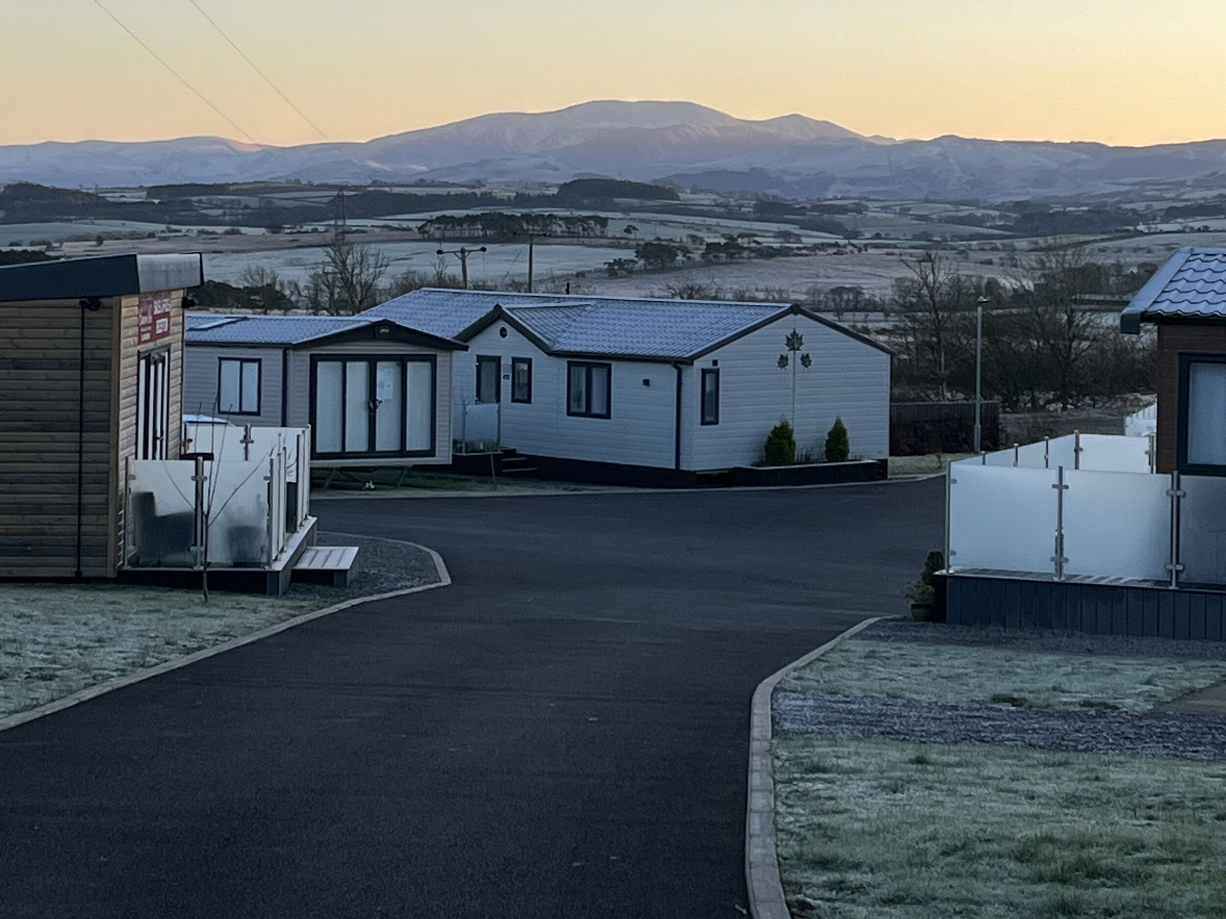 A row of mobile homes with a paved driveway leading to a grassy area, set against a backdrop of distant mountains.
