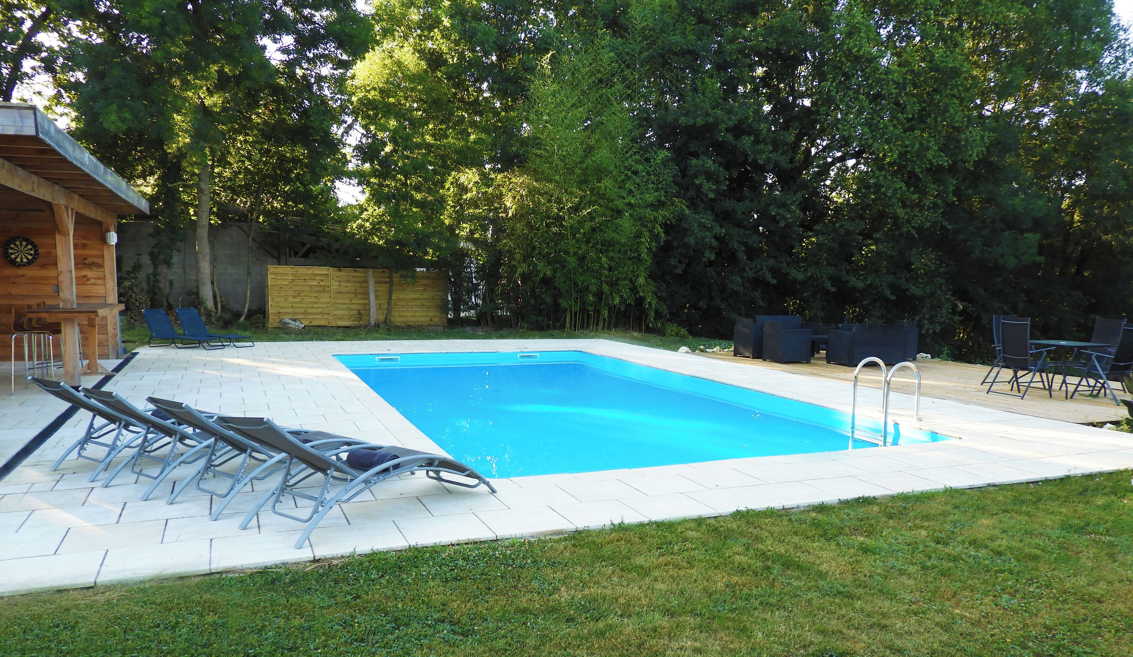 Ein rechteckiger Pool mit einer Metallleiter, umgeben von einer Holzterrasse. Auf der Terrasse befinden sich Liegestühle und ein kleiner Tisch.