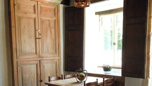 Een rustieke eethoek met een houten tafel en stoelen, een hanglamp en zicht op groen buiten door een raam.