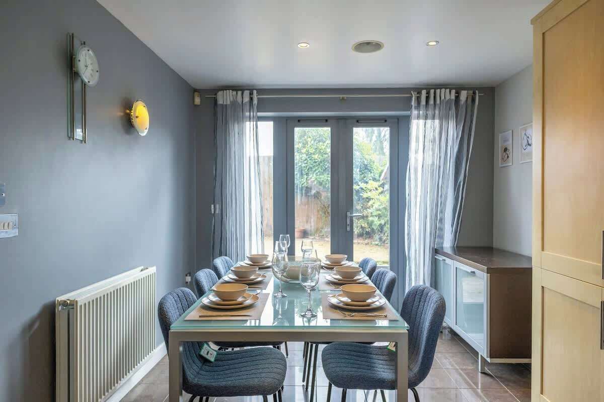 A dining area with a table set for six, a radiator, a clock, and a view of the outdoors.