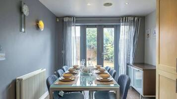A dining area with a table set for six, a radiator, a clock, and a view of the outdoors.