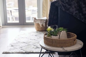 A wicker basket with plants on a small round table, a dark sofa, and a view of outdoor seating through a glass door.