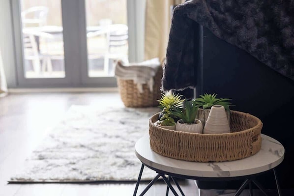 A wicker basket with plants on a small round table, a dark sofa, and a view of outdoor seating through a glass door.