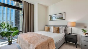 Une chambre d’hôtel moderne avec un grand lit, une table de chevet avec une lampe, une petite plante et une vue sur la ville grâce à de grandes fenêtres.