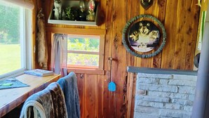 A rustic cabin interior with a wooden ceiling, a stone fireplace, a dining table with a blanket, a rug, and a window with curtains. - Hunting Cabin Rustic Handcrafted primitive sits in our unique rustic Cowboy Town (Pittsford)