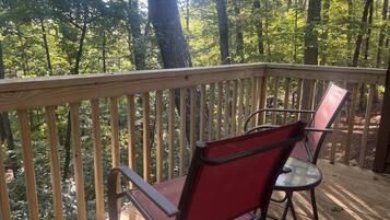Une terrasse en bois avec deux chaises rouges et une table en verre, entourée d’une forêt.