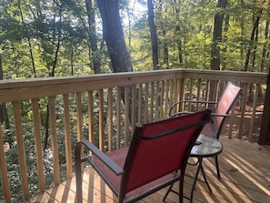 A wooden deck with two red chairs and a glass table, surrounded by a forest.