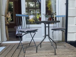 Una terraza de madera con una mesa metálica y dos sillas, una mesita con un jarrón y una cafetera, y vista a zonas verdes a través de una ventana.