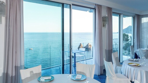 Une chambre dotée de grandes fenêtres donnant sur la mer, un coin repas avec des tables préparées pour deux personnes, et un balcon avec une table et des chaises.