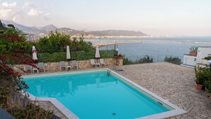 Une piscine rectangulaire bordée de pierres, entourée de chaises longues et de parasols, offrant une vue sur un paysage urbain côtier avec des montagnes à l’horizon.