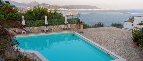 Une piscine rectangulaire bordée de pierres, entourée de chaises longues et de parasols, offrant une vue sur un paysage urbain côtier avec des montagnes à l’horizon.