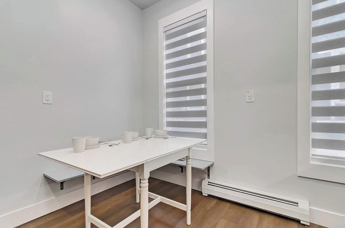 Una habitación con una mesa blanca, dos calentadores fijos en la pared y una ventana con persianas.