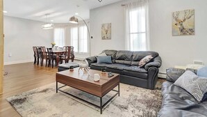 A modern living room with a sofa, coffee table, and dining area. - 4BR Home | Walk Downtown Rutland + Trails (Rutland)