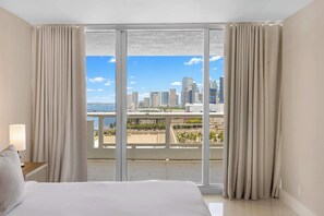 Ein Hotelzimmer mit einem großen Bett, einem Nachttisch und Blick auf eine Stadtlandschaft durch raumhohe Fenster.