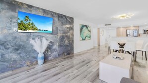 A modern hotel room with a marble wall, a flat-screen TV, a vase with white feathers, a dining area with white furniture, and a kitchen area.