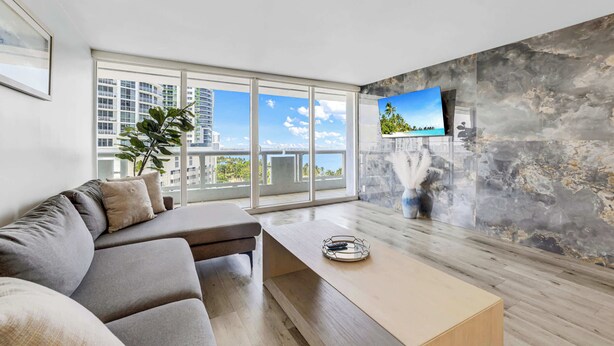 A modern living room with a sofa, a coffee table, a TV mounted on a textured wall, and large windows with a city view.