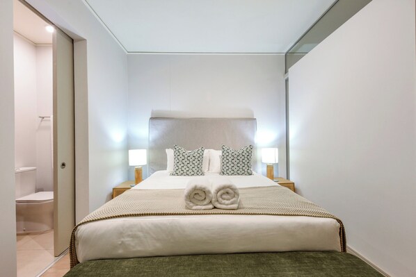 A hotel room with a bed, two bedside lamps, a headboard, and a pair of towels on the bed.