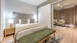 Une chambre moderne avec un grand lit, des tables de chevet, un banc et une vue sur un espace de vie comprenant un canapé et des rideaux.
