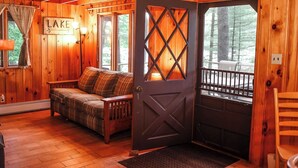 A cabin interior with a plaid sofa, a wooden door with a cross design, and a sign that reads "LAKE". - Gramps Camp • Moose Pond (Bridgton)
