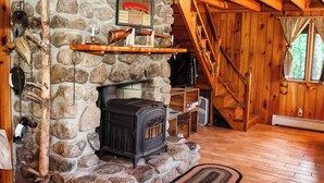 A stone fireplace with a wood-burning stove, a wooden shelf above it, and a stone wall. - Gramps Camp • Moose Pond (Bridgton)